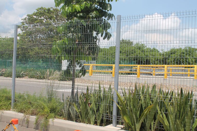 Cerca perimetral de panel metálico galvanizado junto a carril amarillo, plantas y vegetación